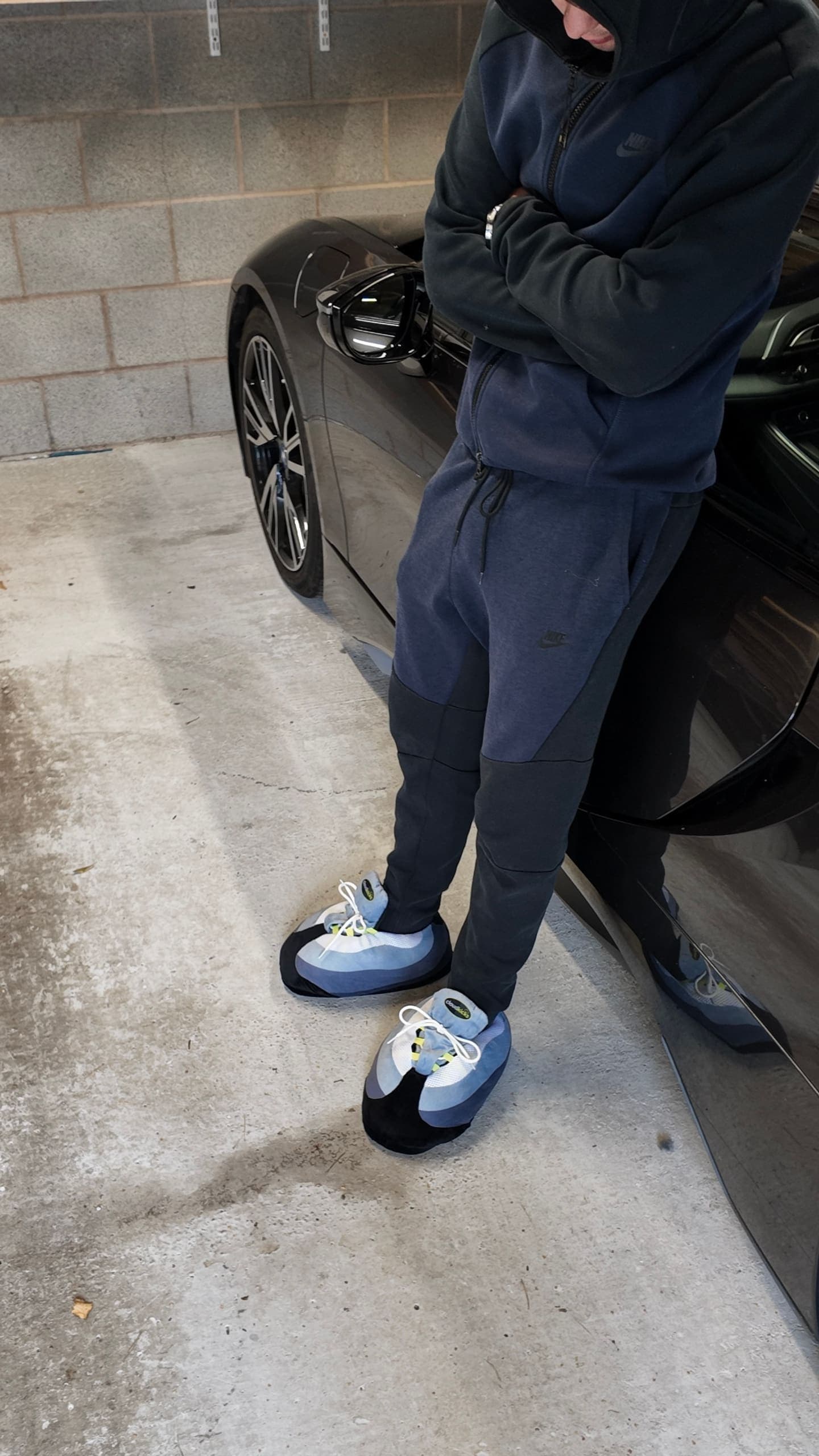 Arms crossed, leaning on a black sports car in a navy tracksuit and OG Neon CloudKicks